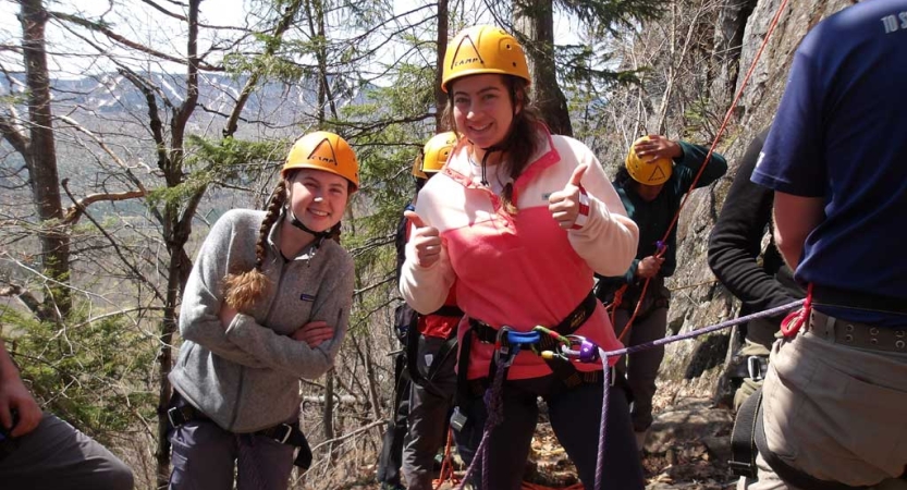 Two people wearing yellow helmets and other safety gear smile at the camera while preparing to rock climb. One of them gives two thumbs up. 